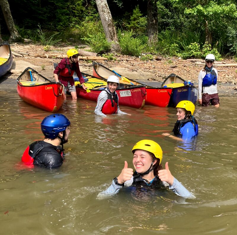 The image shows a group of people in a body of water, likely a lake or river, with canoes nearby. Most of them are wearing helmets and life jackets, suggesting they are participating in water sports activities like canoeing or kayaking. One person in the foreground is giving a thumbs-up. The background features trees and a shoreline, indicating a natural outdoor setting.